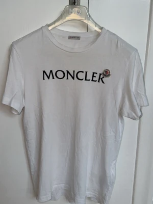 Vit Moncler t-shirt med logga - Vit t-shirt från Moncler med svart tryckt logga på bröstet och broderad Moncler-patch. Klassisk rund halsringning och korta ärmar. Perfekt för dig som gillar stilrena och exklusiva plagg.