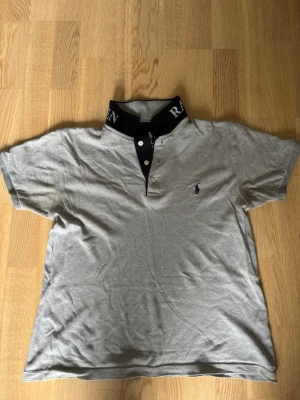 Grå piké från Ralph Lauren - Snygg grå pikétröja från Ralph Lauren med svart krage där märkesnamnet står broderat i vitt. Klassisk passform med korta ärmar och knappslå framtill. Liten logga broderad på bröstet. Perfekt till jeans eller shorts. Personen på bilden är 180, 72 kg, passar mer som S/M. Hör av er vid frågor!