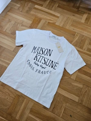 Maison Kitsuné vit t-shirt med tryck - Vit t-shirt från Maison Kitsuné med svart tryck framtill där det står 'Maison Kitsuné Paris, France'. Klassisk passform och rund hals. Tillverkad i mjuk bomull och har en enkel, stilren look som passar till det mesta.