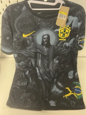Brasilien Nike matchtröja svart - Snygg svart Brasilien matchtröja från Nike med unikt grafiskt tryck framtill, bland annat Kristusstatyn och brasilianska symboler. Gula detaljer med Nike-logga och Brasilien-emblem på bröstet. Korta ärmar och lätt, sportigt material.