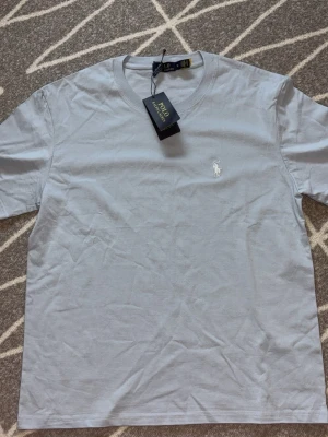Ljusblå t-shirt från Polo Ralph Lauren - Snygg ljusblå t-shirt från Polo Ralph Lauren med klassisk passform och korta ärmar. T-shirten har en broderad vit logga på bröstet och är tillverkad i mjuk bomull. Perfekt för en clean och stilren look.