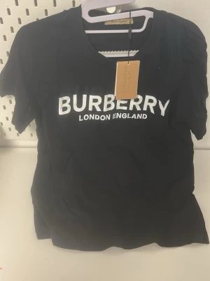 Svart Burberry t-shirt med logga - Svart t-shirt från Burberry med vit logga 'BURBERRY LONDON ENGLAND' tryckt på bröstet. Klassisk rund hals och korta ärmar. Tillverkad i mjuk bomull som känns skön mot huden. Perfekt för en stilren och trendig look.