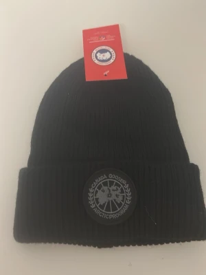Svart mössa från Canada Goose - Svart ribbstickad mössa från Canada Goose med uppvikt kant och rund logotyp-patch framtill. Klassisk och stilren design som passar perfekt till kalla dagar.