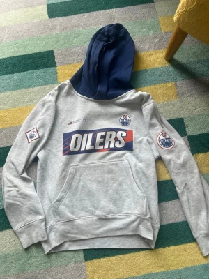 Grå NHL Oilers hoodie med blå huva - Snygg grå hoodie från NHL med Edmonton Oilers-tryck på bröstet och loggor på ärmarna. Mörkblå huva och stor magficka framtill. Perfekt för dig som gillar hockey och vill ha en sportig stil.