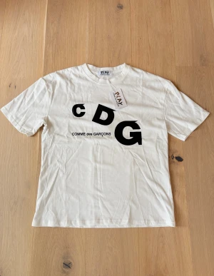 Vit CDG t-shirt från Comme des Garçons - Vit t-shirt från Comme des Garçons PLAY med stor svart CDG-logga framtill. Klassisk passform med rund hals och korta ärmar. Tillverkad i mjuk bomull som känns skön mot huden. Perfekt för dig som gillar stilrena och trendiga plagg.
