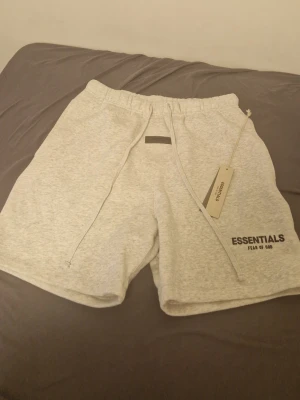 Grå Essentials Fear of God shorts - Säljer ett par ljusgrå shorts från Essentials Fear of God med snörning i midjan och tryckt logga på vänster ben. Sköna och stilrena, perfekta för sommaren. Tillverkade i mjuk bomullsmix och har en relaxed passform.
