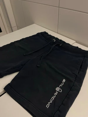 Mörkblå shorts från Sail Racing - Mörkblå shorts från Sail Racing med broderad logga på ena benet. De har snörning i midjan och är tillverkade i mjukt bomullsmaterial. Perfekta för sommardagar och chill stunder.