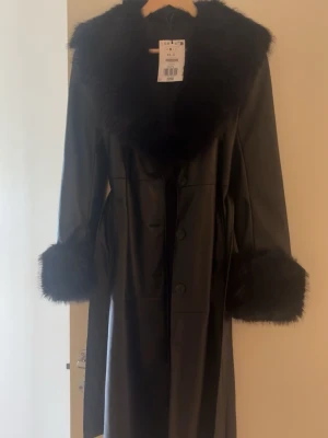 Bershka kappa - Stilren svart lång kappa i skinnimitation med bred krage och muddar i mjuk fuskpäls. Kappan har knäppning framtill och ett matchande skärp i midjan för en snygg siluett💕 kappan är i nytt skick den är helt oanvänd och har även prislappen kvar 
