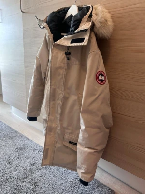 Beige parkas från Canada Goose - Snygg beige parkas från Canada Goose med klassisk röd logotyp på ärmen. Jackan har en stor huva med pälsdetalj, svarta muddar och flera praktiska fickor. Perfekt för kalla dagar tack vare det tjocka fodret och vindtäta materialet. Man kan fälla fram luvan så den täcker hela ansiktet vid kallt väder. Säljer pga ingen användning av längre. OBS! Bild på tags finns, men har slarvat bort så följer inte med i paketet.