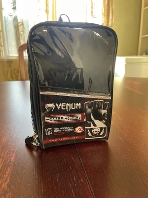 Venum MMA handskar Challenger L/XL - Säljer ett par helt nya svarta Venum Challenger MMA-handskar i storlek L/XL. Handskarna är tillverkade av syntetiskt läder med förstärkta sömmar och lager av skum för extra skydd och stötdämpning. Kardborreband för säker passform. Perfekta för träning och matcher! 