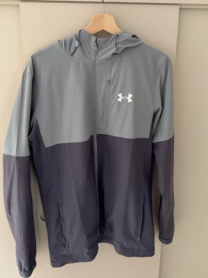Grå och mörkblå vindjacka Under Armour - Snygg tvåfärgad vindjacka från Under Armour i grått och mörkblått. Jackan har huva, dragkedja framtill, bröstficka med dragkedja och två sidofickor. Perfekt för blåsiga dagar och sportiga outfits. Under Armour-logga på bröstet.