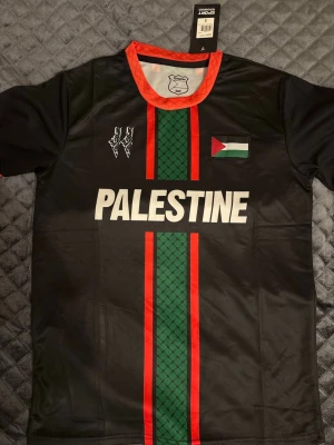 Svart Palestine fotbollströja S - Snygg svart fotbollströja från PC Palestina med röd och grön rand i mitten, vit text 'PALESTINE' och palestinska flaggan på bröstet. Röd krage och sportig design i funktionsmaterial. Perfekt för dig som vill visa ditt stöd på planen eller läktaren.