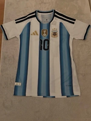 Argentina Messi matchtröja Adidas - Säljer en officiell Argentina fotbollströja från Adidas med Messi och nummer 10 på ryggen. Tröjan har klassiska blå och vita ränder, svarta detaljer på axlarna och broderade märken på bröstet. Perfekt för dig som älskar fotboll och vill stötta ditt favoritlag.