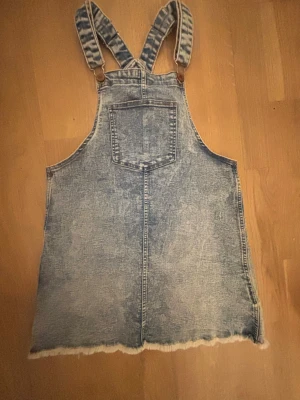 Ljusblå jeansdress med hängslen - Säljer en ljusblå jeansdress i klassisk denim med justerbara axelband och en stor bröstficka. Rå kant nertill för en avslappnad vibe. 
