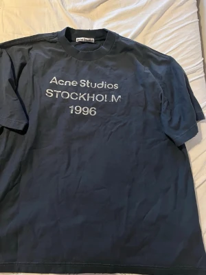 Blå Acne Studios t-shirt Stockholm - Storlek s men passar mer som mSnygg mörkblå t-shirt från Acne Studios med tryck 'STOCKHOLM 1996' på bröstet. Klassisk passform och rund halsringning. Perfekt för dig som gillar stilrena och trendiga plagg med streetkänsla.
