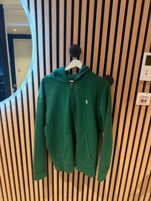 Grön hoodie från Polo Ralph Lauren - Snygg grön hoodie från Polo Ralph Lauren med dragkedja framtill och klassisk vit logga på bröstet. Hoodien har huva, två fickor och ribbade muddar vid ärmar och nederkant. Perfekt för en chill och avslappnad stil. passar xl or L jag är 195 den är lite kort på armarna