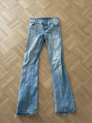 Ljusblå bootcut jeans med rå kant - Säljer ett par ljusblå bootcut jeans med rå och fransig kant längst ner. Värt att veta att jeansen har ett litet hål där bak men som snabbt går att laga. Jeansen har dubbla knappar i midjan, klassiska fickor och markerade sömmar längs benen. Perfekt för dig som gillar en avslappnad och trendig look.