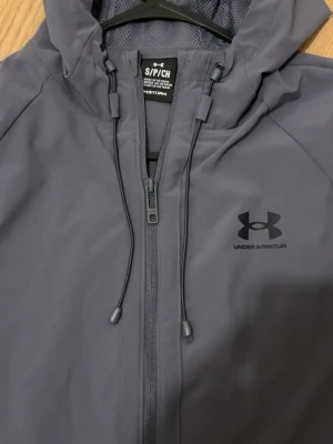 Grå vindjacka från Under Armour - Snygg grå vindjacka från Under Armour med dragkedja framtill, justerbar huva och två sidofickor. Jackan har en minimalistisk design med logga på bröstet och är tillverkad i ett lätt, vattenavvisande material som passar perfekt för blåsiga dagar.