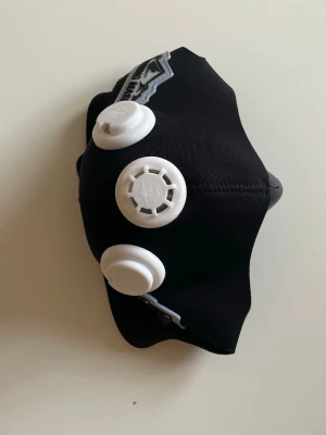 Svart cosplaymask med ventiler - Cool svart maskeradmask från Training Mask med tre vita ventiler framtill. Masken är tillverkad i elastiskt material och har justerbara band för att sitta bekvämt över ansiktet. Perfekt för cosplay eller maskerad där du vill sticka ut.