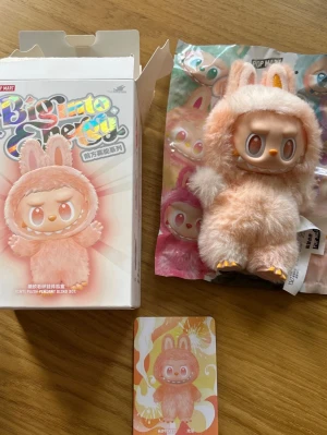 Pop Mart Labubu - Söt samlarfigur från Pop Mart Big Into Energy-serien. Figuren har mjuk, fluffig päls i ljusrosa och beige, med kaninöron och orange detaljer. Kommer med originalförpackning och samlarkort. Perfekt för dig som gillar unika och gulliga prydnader.