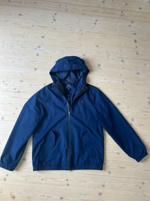 Marinblå vindjacka från Polo Ralph Lauren - Riktigt fer marinblå vindjacka från Polo Ralph Lauren med huva, dragkedja framtill och två fickor med lock. Jackan har elastiska muddar och nederkant samt en liten röd ralph lauren logga broderad på bröstet. Enda ”skavanken” är ett minimalt hål vid insidan av luvan (med på bild.) priset är ej hugget i sten, hör av et vid frågor!