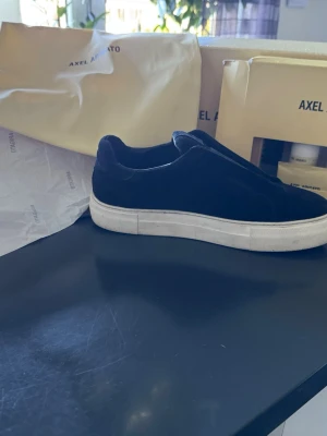 Svarta slip-on sneakers Axel Arigato - Stilrena svarta slip-on sneakers från Axel Arigato med ovandel i mocka och tjock vit gummisula. Minimalistisk design utan snörning, perfekt för en clean och modern look. Kommer med originalkartong och dustbag.  Du får med att klining kitt till skorna .