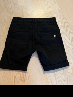 Svarta shorts med nitar och stjärndetalj - Snygga svarta shorts med silverfärgade nitar längs fickorna framtill och en liten stjärndetalj på ena bakfickan. Klassisk femficksmodell med uppvikta benslut och dragkedja. Perfekta för en trendig och avslappnad stil.