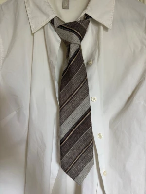 Brun slips med breda ränder - Säljer en stilren slips i brunt med breda diagonala ränder i beige, vitt och mörkbrunt. Perfekt för dig som vill addera lite retrovibe till outfiten.