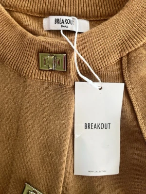Beige kofta från Breakout - Stilren beige kofta från Breakout med raka, vida ärmar och dekorativa metallspännen framtill. Koftan har en mjuk, stickad känsla och rund halsringning. Perfekt för dig som gillar minimalistisk och trendig design.