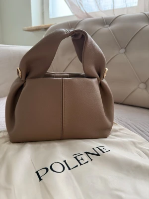 Polène brun handväska i läder - Snygg handväska från Polène i mjukt, brunt läder med unik, knuten design på handtaget. Väskan har en stilren och modern look med guldfärgade detaljer på sidorna. Perfekt accessoar för att lyfta din outfit.