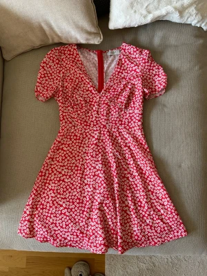 Röd blommig kortklänning Chiquelle - Supersöt röd kortklänning från Chiquelle med vitt blommönster. Klänningen har v-ringning, korta puffärmar och markerad midja. Perfekt för sommarens alla tillfällen! Lätt och luftig känsla.