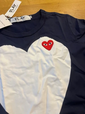 Comme des Garçons Play hjärttshirt - Mörkblå t-shirt från Comme des Garçons Play med stort vitt hjärta på bröstet och rött broderat hjärta med ögon. Klassisk rund hals och korta ärmar. Perfekt statement-plagg för dig som gillar streetstyle.