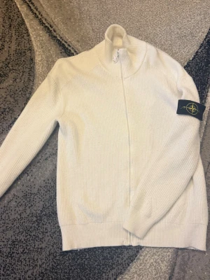 Vit stickad ziptröja Stone Island - Vit stickad ziptröja från Stone Island med hög krage och ribbstickad struktur. Tröjan har dragkedja framtill och den klassiska logotypen på vänster ärm. Perfekt för lager-på-lager och snygg till jeans.