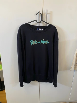 Rick and Morty svart sweatshirt - Svart sweatshirt med Rick and Morty-tryck framtill och en färgglad illustration av Rick på skateboard på ryggen. Tröjan har rund halsringning och långa ärmar. Perfekt för dig som gillar coola och unika motiv från populära serier.