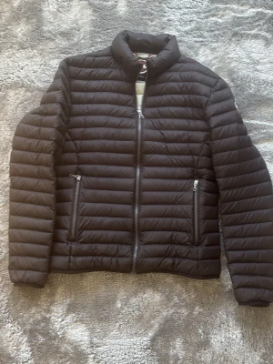 Svart dunjacka från Colmar - Säljer en svart dunjacka från Colmar Originals med quiltad design och två fickor med dragkedja. Jackan har hög krage och snygga detaljer, perfekt för kalla dagar. Materialet är lätt och isolerande, vilket gör den riktigt varm och bekväm.