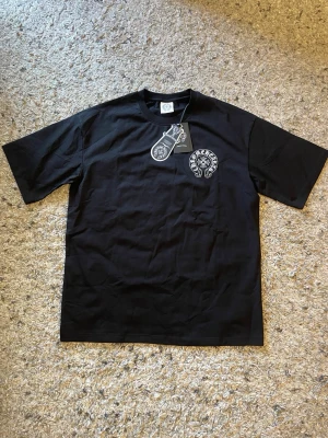 Svart Chrome Hearts t-shirt - Svart t-shirt från Chrome Hearts med vit logga på bröstet och stort tryck på ryggen. Klassisk passform och rund halsringning. Perfekt för dig som gillar streetwear och vill sticka ut med en ikonisk design.