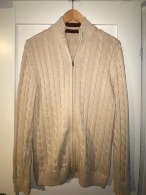 Beige kabelstickad ziptröja - Snygg beige kabelstickad tröja med dragkedja framtill och hög krage. Tröjan har långa ärmar och ribbade muddar. Tröjan är storlek S men med tanke på att den är rättså lång så skulle jag säga att den sitter som en Slim M. Säljer den på grund av att den inte var bra storlek för mig. Tagen vid kragen är av klippt då den var extremt irriterande i nacken.