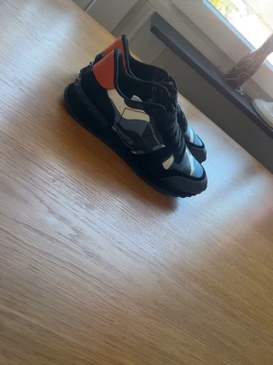 Valentino Garavani camo sneakers - Snygga sneakers från Valentino Garavani med svart, vit och grått camo-mönster samt en röd detalj på hälen. Skorna har svarta snören och en robust sula. Materialmix av läder och textil ger en exklusiv känsla och cool look. Skorna är helt nya och oanvända men fel storlek för mig. Vill sälja snabbt så öppen för bud!