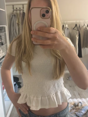 Vit smockad topp med volang - Säljer en söt vit topp med smockad midja och volang nertill. Toppen är kortärmad och har en feminin, figurnära passform. Perfekt att styla med jeans eller kjol för en trendig look. 👌👌