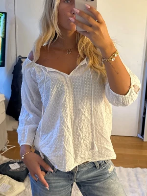 Vit offshoulder blus med struktur - Snygg vit blus med offshoulder-modell och trekvartsärmar. Blusen har en strukturerad yta med diskret rutmönster och knytband vid halsen. 