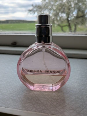 Ariana Grande parfym Cloud pink  - Säljer en Ariana Grande parfym i en snygg, rund rosa glasflaska med silvrig spraykork. Flaskan har en modern design och är genomskinlig med rosa ton. Perfekt för dig som gillar kändisparfymer och vill ha något fräscht och trendigt i samlingen. Mängd enligt bild. Tyvärr saknas korken. 