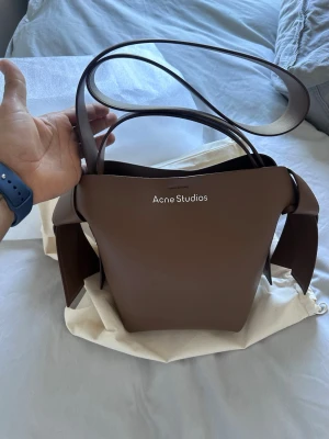 Brun handväska Acne Studios - Snygg brun handväska från Acne Studios i slät läder med minimalistisk design och bred axelrem. Väskan har en unik form med vikta sidor och logga framtill. Medföljer dustbag i beige tyg. Perfekt för dig som gillar stilrena accessoarer.