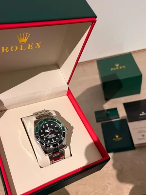 Role Submariner med grön bezel - Säljer en stilren Role Submariner med svart urtavla och grön bezel. Klockan har ett klassiskt armband i rostfritt stål och levereras i originalbox med certifikat och tillbehör. Perfekt för dig som gillar exklusiva accessoarer och tidlös design.