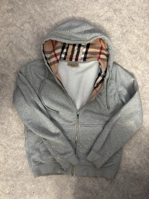 Grå zip hoodie från Burberry - Snygg grå zip hoodie från Burberry med klassiskt rutigt foder i huvan. Hoodien har dragkedja framtill, två fickor och ribbade muddar vid ärmar och nederkant. Tillverkad i mjukt bomullsmaterial och har justerbar dragsko i huvan.