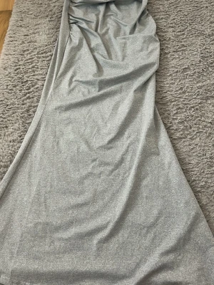 Lång silverklänning med tunna band - Snygg långklänning i silver med tunna axelband och draperad urringning. Klänningen har en slät, glittrig yta och faller mjukt längs kroppen. Perfekt för dig som vill sticka ut med en elegant look.