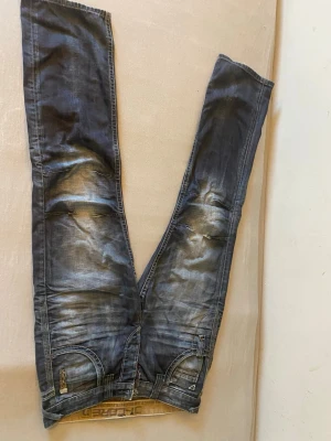 Blå slitna jeans med detaljer - Säljer ett par blå jeans med slitningar och tvättade effekter. Jeansen har raka ben, tydliga sömmar och coola detaljer vid fickorna. Materialet är klassisk denim och de har en avslappnad streetstil som passar perfekt till vardagslooken.
