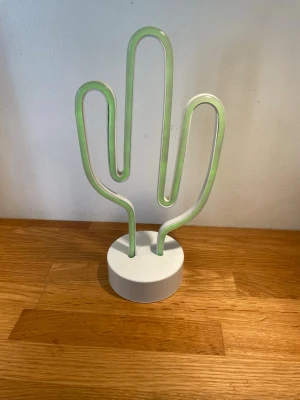 Neonlampa kaktusformad med vit fot - Cool neonlampa i form av en kaktus med vit rund fot. Lampan har en böjd design och lyser i grönt när den är påslagen, annars är den vit. Perfekt som dekorativ belysning i rummet. Materialet är plast och lampan är lätt att placera på en hylla eller ett bord.