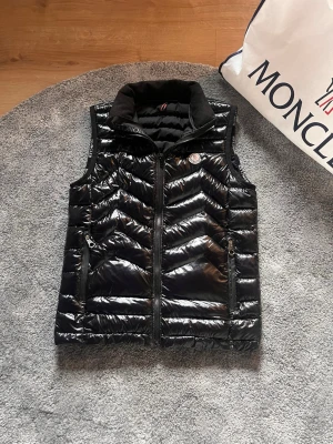 Svart dunväst  - Snygg svart dunväst från Moncler med glansig finish och klassisk Moncler-logga på bröstet. Västen har hög krage i mjukt material, dragkedja framtill och två fickor med dragkedja. Perfekt för lager på lager och streetstyle. Fodral på bild följer ej med