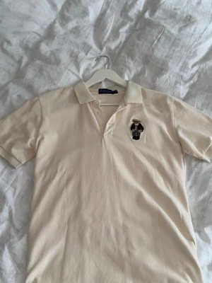 Beige pikétröja från Ralph Lauren Purple Label - Klassisk beige pikétröja från Ralph Lauren med broderad nalle på bröstet. Tröjan har krage, korta ärmar och knäppning framtill. Perfekt för en avslappnad men stilren look.