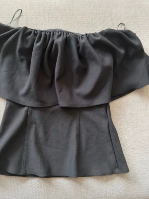 Svart offshoulder topp med volang - Snygg svart offshoulder topp med bred volang runt bröstet och axelbandslös design. Toppen har en figurnära passform och är perfekt för dig som vill ha en trendig och stilren look. Tillverkad i mjukt material som sitter skönt mot kroppen.